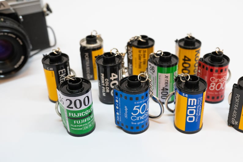 May include: A collection of film roll keychains in various colors, including yellow, black, blue, and red. Each keychain features film brand labels and a metal keyring. A vintage camera is visible in the background.