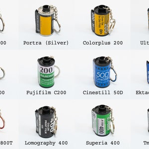 May include: A collection of miniature film roll keychains. The keychains feature various film brands and types, including Portra 400, Ektar 100, and Fujifilm C200. Each keychain has a silver keyring attached.