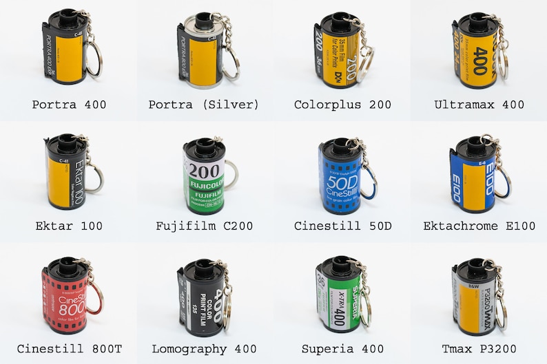 May include: A collection of miniature film roll keychains. The keychains feature various film brands and types, including Portra 400, Ektar 100, and Fujifilm C200. Each keychain has a silver keyring attached.