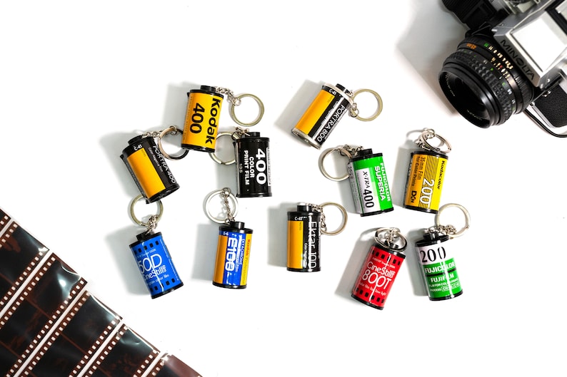 May include: A collection of film roll keychains in various colors, including yellow, black, red, blue, and green. The keychains feature text such as "Kodak 400", "Ektar 100", and "Fujicolor 200". A vintage camera is visible in the background.