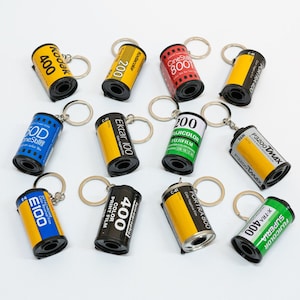 May include: A collection of film canister keychains in various colors, including yellow, red, blue, and black. Each keychain features film brand names and numbers like Kodak 400, Ektar 100, and Fujifilm. The keychains are arranged on a white surface.