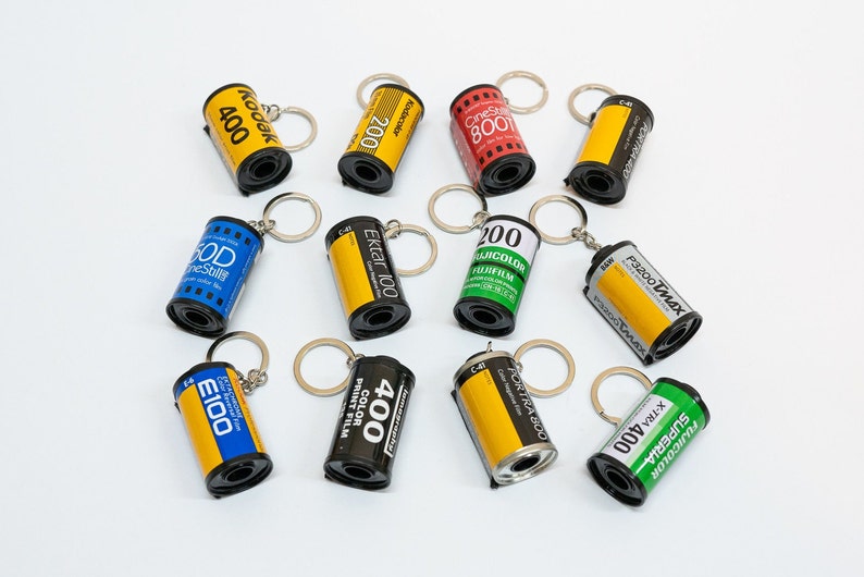 May include: A collection of film canister keychains in various colors, including yellow, red, blue, and black. Each keychain features film brand names and numbers like Kodak 400, Ektar 100, and Fujifilm. The keychains are arranged on a white surface.