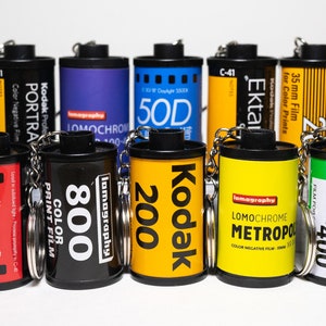 May include: A collection of film canister keychains in various colors, including black, yellow, and blue. Each keychain features film brand names and specifications, such as Kodak, Lomography, and Fujifilm. The keychains are arranged on a white surface.