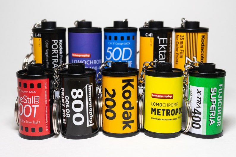 May include: A collection of film canister keychains in various colors, including black, yellow, and blue. Each keychain features film brand names and specifications, such as Kodak, Lomography, and Fujifilm. The keychains are arranged on a white surface.