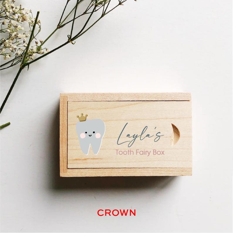 May include: A wooden tooth fairy box with a light gray tooth with a gold crown and a pink blush on its cheeks. The box has the text "Layla's Tooth Fairy Box" on it.