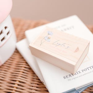 May include: A wooden box with a floral design and the text "Layla's Keepsakes" on the top. The box is sitting on top of a stack of books.