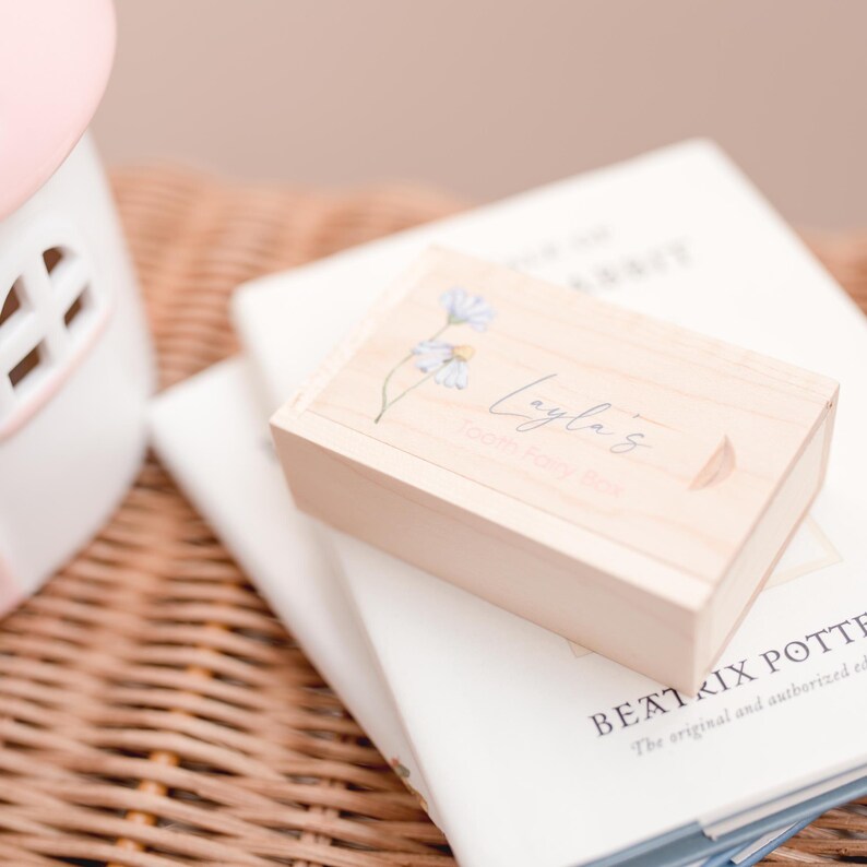 May include: A wooden box with a floral design and the text "Layla's Keepsakes" on the top. The box is sitting on top of a stack of books.