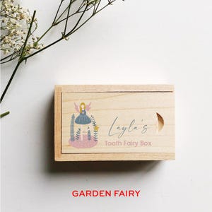 May include: A wooden box with a light wood finish. The box has a hinged lid and a cutout for a handle. The lid is decorated with a colorful illustration of a fairy with wings, a pink heart, and green leaves. The text "Layla's Tooth Fairy Box" is printed on the lid.