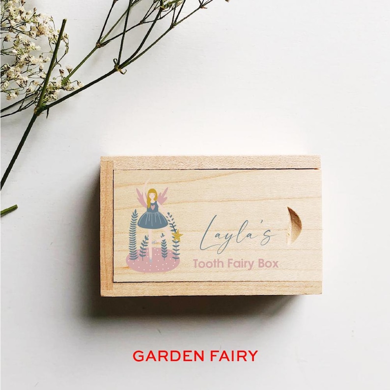 May include: A wooden box with a light wood finish. The box has a hinged lid and a cutout for a handle. The lid is decorated with a colorful illustration of a fairy with wings, a pink heart, and green leaves. The text "Layla's Tooth Fairy Box" is printed on the lid.