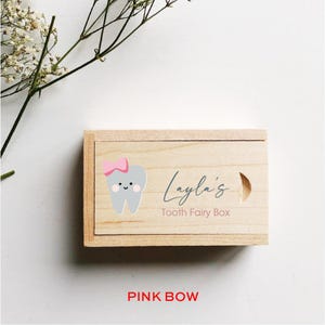 May include: A wooden tooth fairy box with a light wood finish. The box has a gray tooth with a pink bow and a smiling face on the front. The text "Layla's Tooth Fairy Box" is printed on the box.