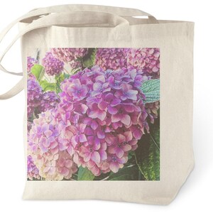Puede incluir: Un bolso tote beige con una imagen de primer plano de flores de hortensia rosas y moradas. Las flores tienen un aspecto suave y vintage con hojas verdes. El bolso tiene dos asas.