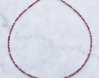 Op de afbeelding: Een delicate gouden ketting met kleine, donkerrode kralen. De ketting is gemaakt van goudgevulde draad en heeft een eenvoudige sluiting.