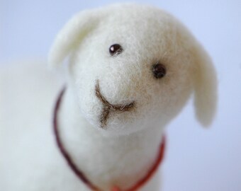 May include: A white felt sheep with brown eyes and a stitched smile, wearing a red and brown cord necklace with a silver bell charm.