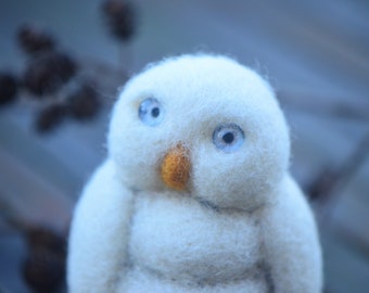 May include: A white, fluffy, felt owl with blue eyes and an orange beak. The owl is sitting on a wooden surface.