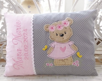 Puede incluir: Una almohada gris y rosa con un patrón de cuadros rosa y un borde blanco festoneado. La almohada tiene un nombre bordado en rosa "Mara Marie" y la fecha "20.02.2024". El otro lado de la almohada presenta un oso de peluche marrón que lleva un vestido de cuadros rosa con un corazón rosa y una corona de flores rosas.