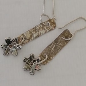 May include: A pair of silver earrings with a textured rectangular charm and a small flower charm. The flower charm has a small pink stone in the center.