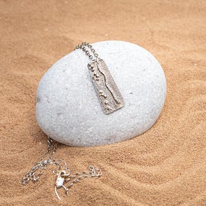 Peut inclure: Un collier en argent avec un pendentif rectangulaire texturé présentant un motif de ligne ondulée en relief. Le collier est présenté sur une pierre gris clair sur un fond de sable.
