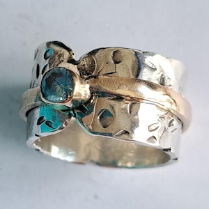 May include: A silver and gold ring with a blue gemstone set in the center. The ring is textured and has a unique design.