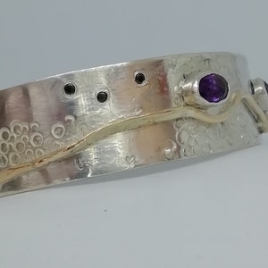 Op de afbeelding: Zilveren en gouden armband met een getextureerd oppervlak en twee paarse edelstenen. De armband heeft een golvend gouden band met een getextureerd oppervlak.