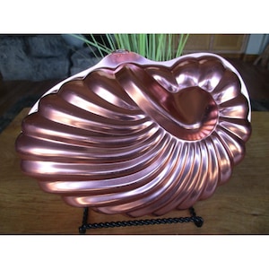 May include: A copper-coloured, nautilus-shaped bundt pan. The pan features a detailed, ridged design and rests on a black metal stand. The pan's surface reflects light, highlighting its curves and intricate details. The overall theme is kitchenware.