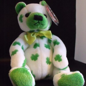 May include: White plush bear with green shamrock print and a green bow tie. The bear has a Ty tag attached to its ear.