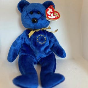 May include: A plush blue teddy bear with a gold ribbon around its neck and a patch of yellow stars. The bear has a red and white tag attached to its ear. The bear is a collectible item.