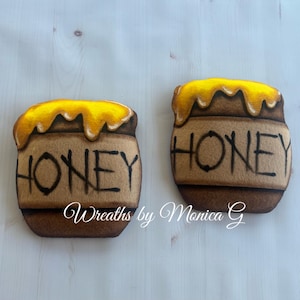 May include: Two felt honey pot decorations. Each pot is brown with the word "HONEY" in black. The top of each pot has a yellow felt honey spill. The bottom of the image has the text "Wreaths by Monica G".