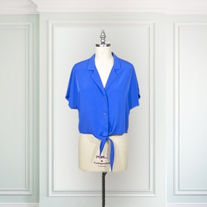 May include: A blue silk blouse with a tie front and short sleeves. The blouse is on a white dress form.