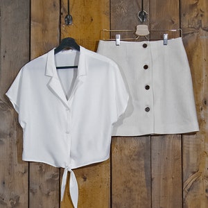 May include: A white button-down shirt with short sleeves and a tie at the waist, and a beige linen skirt with a button closure.