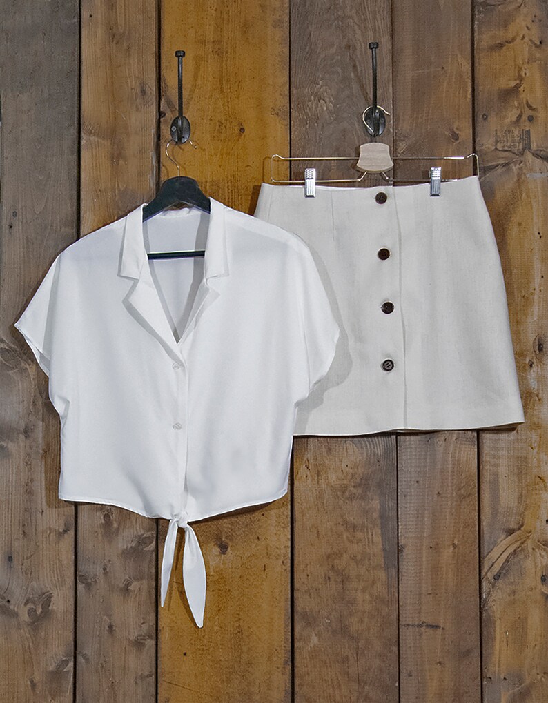 May include: A white button-down shirt with short sleeves and a tie at the waist, and a beige linen skirt with a button closure.