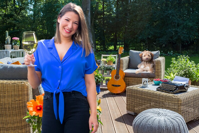 May include: A woman in a blue shirt and black pants is holding a glass of white wine. She is standing on a wooden deck with a wicker chair, a guitar, and a dog in the background.
