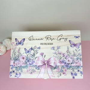 May include: A light-colored wooden box with a floral and butterfly design. The box features a large pink bow, purple flowers, and butterflies. The text "Eleanor Rose Grey" and the date "05/08/2025" are printed on the box.