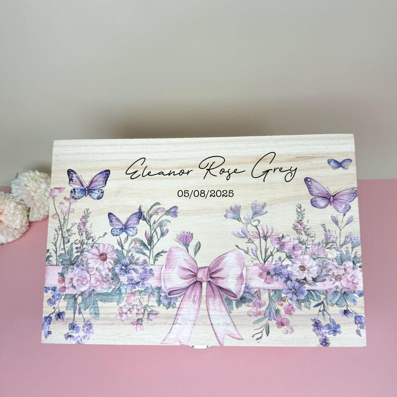 May include: A light-colored wooden box with a floral and butterfly design. The box features a large pink bow, purple flowers, and butterflies. The text "Eleanor Rose Grey" and the date "05/08/2025" are printed on the box.