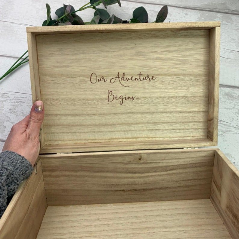 May include: A wooden box with a hinged lid. The lid is open and the inside of the box is visible. The inside of the box is made of wood and has the words "Our Adventure Begins" engraved on it.