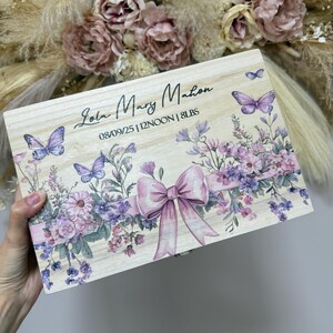 May include: Wooden keepsake box with a floral and butterfly design. The box features a large pink bow and the name "Lola Mary Mahon" along with the date, time, and weight. The box is decorated with purple and pink flowers and butterflies.