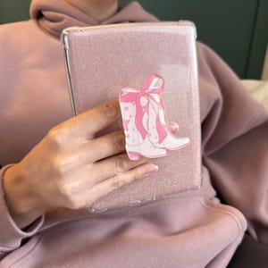 Könnte beinhalten: Eine rosa glitzernde Handyhülle mit einer transparenten Überlagerung, die eine rosa und weiße Illustration von Cowboystiefeln mit einer rosa Schleife zeigt.