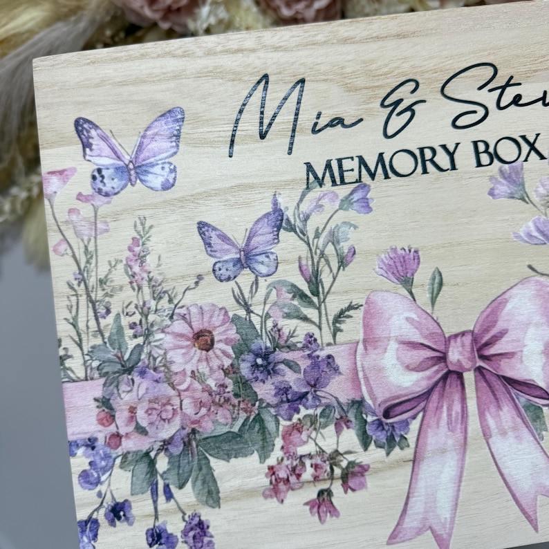 May include: Wooden memory box with floral and butterfly design. The box features a light wood grain finish, with a watercolor-style floral arrangement in shades of pink, purple, and green. The text "Mia & Ste Memory Box" is printed on the box.
