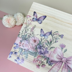May include: Wooden box with a floral and butterfly design. The box features a light wood grain finish, with a watercolor-style illustration of purple butterflies, flowers, and a pink bow. The design is on the top of the box.
