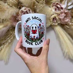May include: White ceramic mug with a wraparound design featuring a cartoon ghost reading a book, surrounded by colourful flowers. The text "ANTI SOCIAL BOOK CLUB" is displayed around the design. The mug has a curved handle.