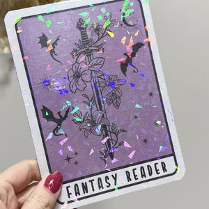 May include: A purple tarot card with a holographic finish. The card features a black and white illustration of a sword with flowers and bats. The text "FANTASY READER" is printed on the card.