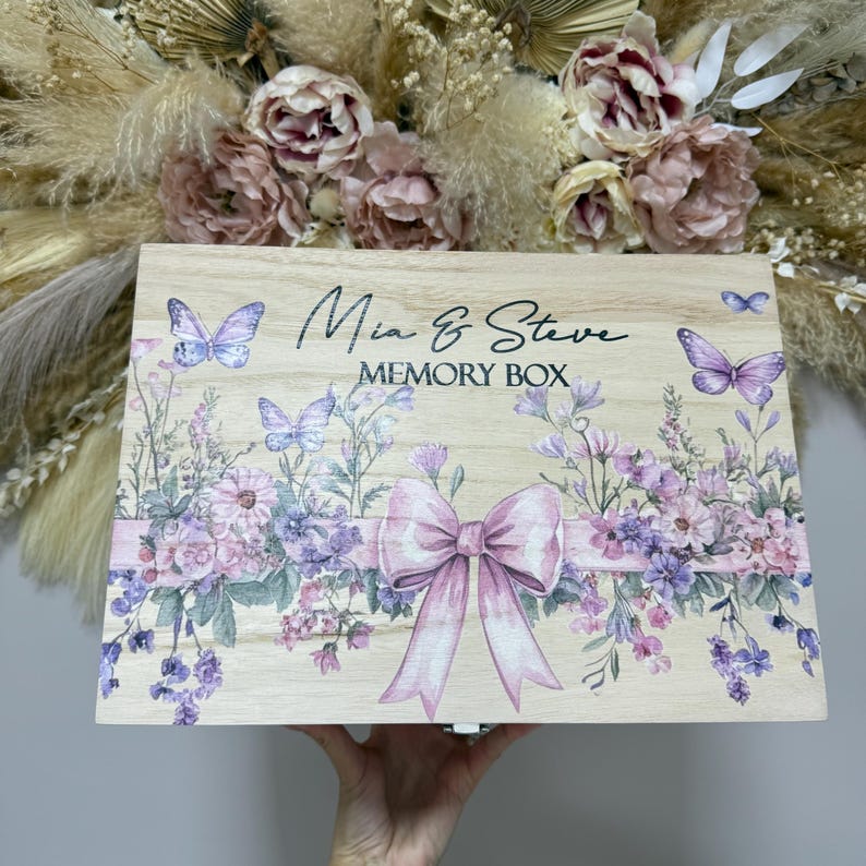 May include: A wooden memory box with the names "Mia & Steve" and the words "Memory Box" printed on the lid. The box is decorated with watercolor-style butterflies, flowers, and a pink bow. The background features dried flowers.