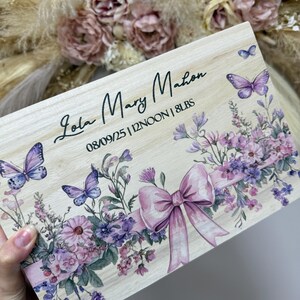 May include: A wooden keepsake box with the name "Lola Mary Mahon" and birth details. The box features a floral design with purple butterflies and a pink bow. The birth weight is listed as 8 lbs.