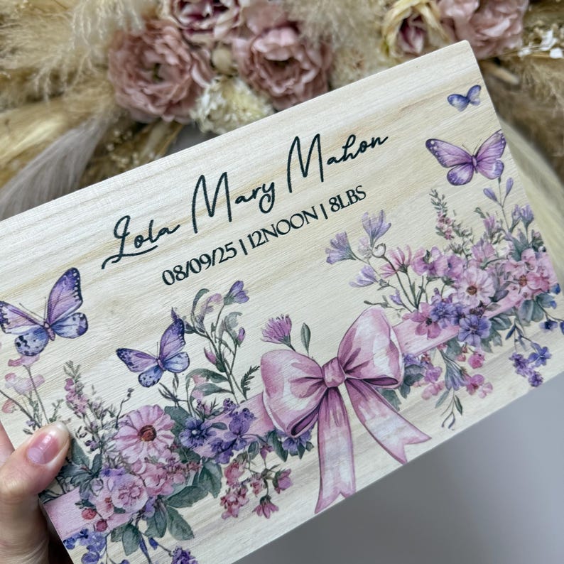 May include: A wooden keepsake box with the name "Lola Mary Mahon" and birth details. The box features a floral design with purple butterflies and a pink bow. The birth weight is listed as 8 lbs.