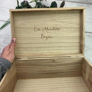 May include: Open wooden box with the words "Our Adventure Begins" engraved on the inside of the lid. The box is light brown and appears to be made of wood. A hand is holding the box open. Greenery is visible in the background.