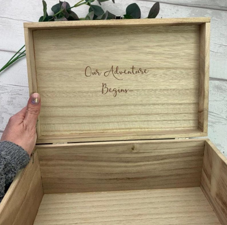 May include: Open wooden box with the words "Our Adventure Begins" engraved on the inside of the lid. The box is light brown and appears to be made of wood. A hand is holding the box open. Greenery is visible in the background.