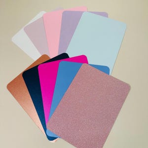 Könnte beinhalten: Eine Sammlung von zehn verschiedenen farbigen Kartonbögen. Die Farben umfassen Weiß, Hellrosa, Lavendel, Hellblau, Kupfer, Dunkelblau, Magenta, Hellblau und zwei Rosatöne.