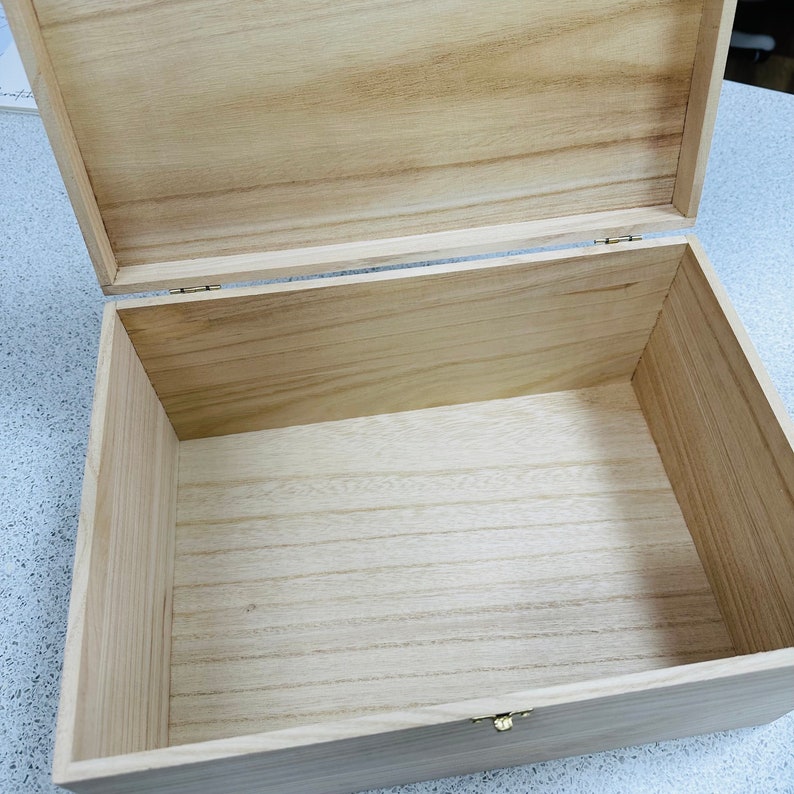 May include: An open, rectangular wooden box with a hinged lid. The box is made of light-colored wood, with visible grain patterns. A small, gold-colored clasp secures the lid. The interior of the box is empty.