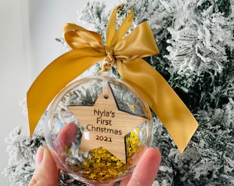 May include: Clear Christmas ornament with a wooden star-shaped insert. The star has the text "Nyla's First Christmas 2021". The ornament is filled with gold star-shaped confetti and has a gold ribbon for hanging.