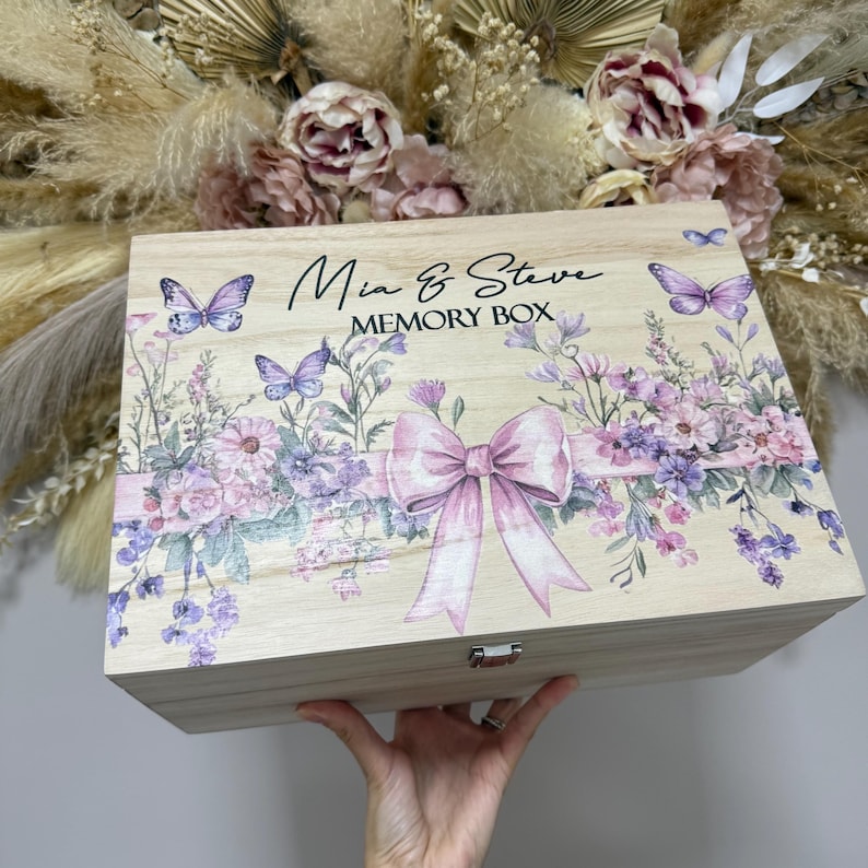 May include: A wooden memory box with a floral design and the text "Mia & Steve MEMORY BOX". The box features a large pink bow, purple butterflies, and lavender flowers. The box is held by a person.
