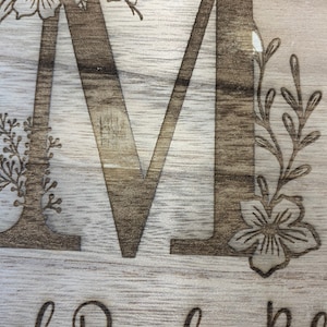 May include: A wooden sign with the letter "M" engraved in the center. The letter is surrounded by floral designs, including a flower and leaves.